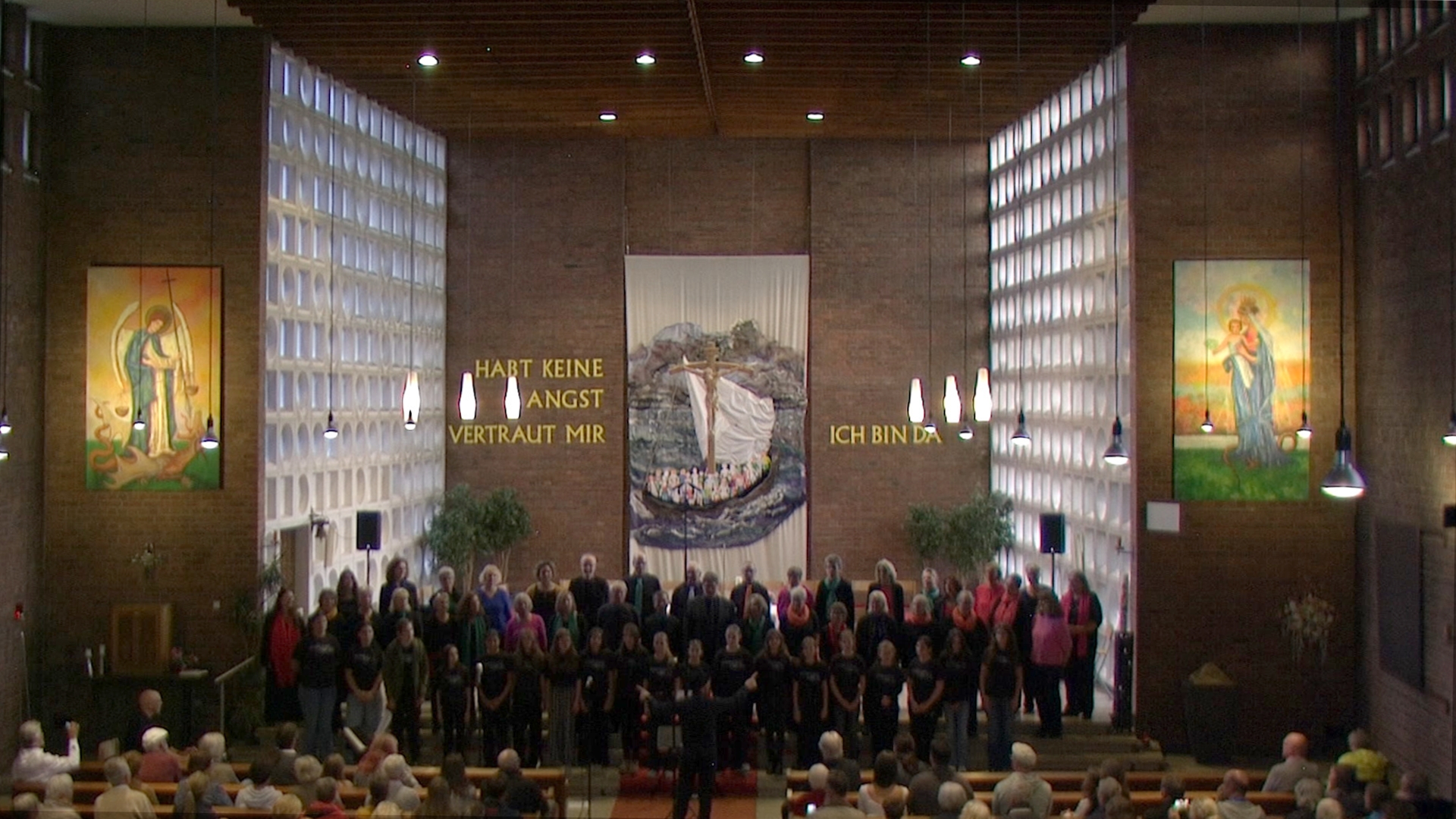 So_soll_es_bleiben-Konzertbild Das Bild zeigt den Kirchraum der Kirche St. Michael in Krefeld Lindental. Zu sehen ist der MEGels-Chor bei seinem Jubiläumskonzert. Gekleidet in Schwarz mit jeweils einem bunten Akzent. Davor steht der Kinderchor "Neverending Voices" des Michael-Endy-Gymnasiums Tönisvorst/NRW. Diese sind in schwarz gekleidet. Der Chorname steht auf den T-Shirts. Im Hintergrund sieht man verschiedene kirchliche Abbildungen und der Text "Habt keine Angst, vertraut mir, Ich bin da".